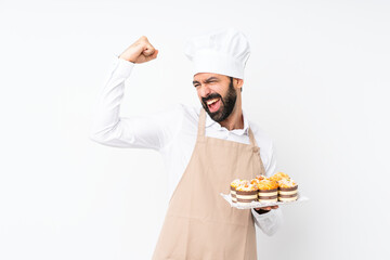 Young man holding muffin cake over isolated white background celebrating a victory