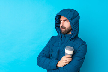 Man wearing winter jacket and holding a takeaway coffee over isolated blue background with confuse...