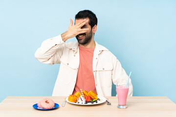 Man at a table having breakfast waffles and a milkshake covering eyes by hands and smiling