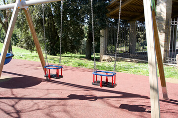 Two empty toddler bucket swings with blue and red seats hang from a wooden frame on a red rubber...