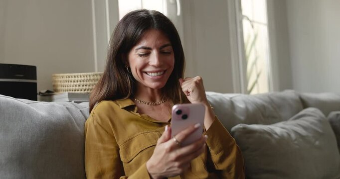Attractive Latina woman sitting on sofa with smartphone, read messages, feels happy about great news in e-mail or sms, get refund, invitation on event, enjoy moment of auction victory, sell-out notice