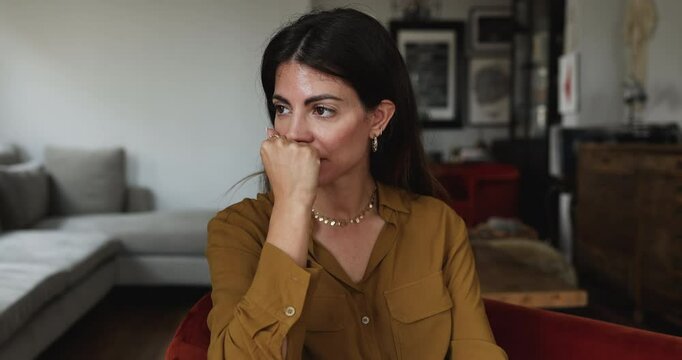 Young pretty Hispanic woman sitting in armchair looks sad while thinking about problem, personal life concerns or unpleasant situation, going though break up, feeling lonely and unhappy, close up shot