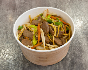 Beef lo mein in a paper bowl with vegetables on a gray surface top