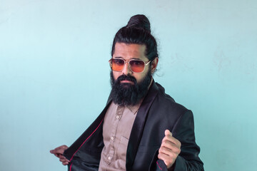 Obraz premium man with long dark hair tied in topknot, full beard, aviator sunglasses. wearing shirt under blazer with red lining. casually adjusting his jacket with one hand and holding his other hand up slightly.
