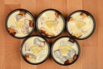 Five small black ramekins contain pieces of biscuit dipped in lemon cream, each garnished with a slice of lemon, arranged on a wooden surface