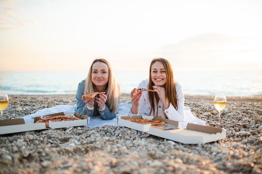 Two girl friends 25-29 year old having picnic with pizza and wine at beach together. Summer vacation season.