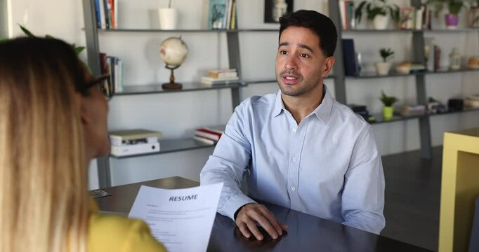 Positive handsome Latin young candidate man speaking to HR agent woman holding paper resume on interview meeting, smiling, telling about work skills, experience