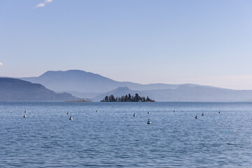 Obraz premium San Biagio island on Lake Garda in Italy surrounded by calm water and distant hills in soft blue morning haze during quiet spring day