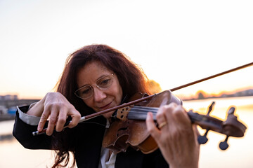 beautiful middle-aged woman dressed in an elegant tailcoat playing a handmade violin at sunset on a small pier