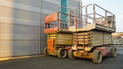 Two parked scissor lifts, one orange and one beige, with their platforms lowered, standing outdoors against a modern building.