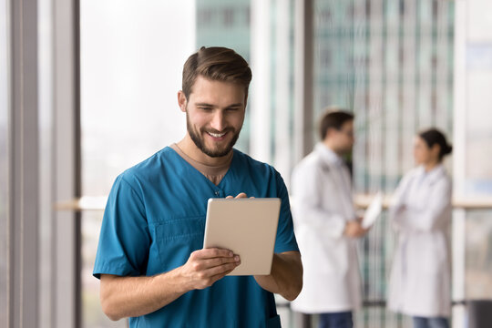 Digital healthcare. Happy smiling young man doctor surgeon use tablet computer at hospital corridor enjoy easy communication with patient on distance read good news in email chat with colleague online
