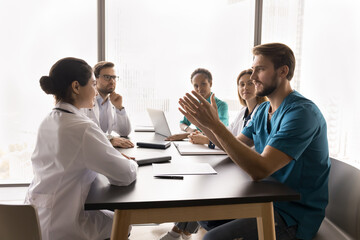 Doctor offer solution. Confident millennial man surgeon speak to multicultural colleagues sitting at table on team briefing explain creative idea propose innovative changes for patient healthcare plan