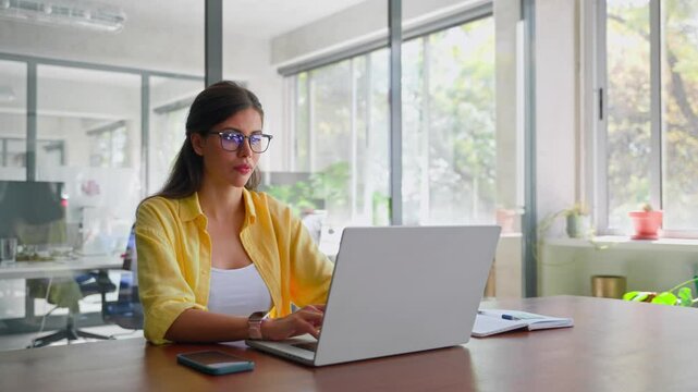 Smiling latin hispanic female marketing manager, professional it specialist working, browsing at laptop computer sitting at desk in modern office. Cheerful young woman employee using pc for business 