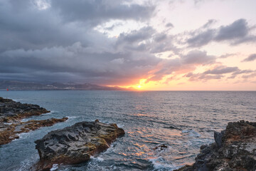Golden hour view of a stunning sunset over the Atlantic Ocean, captured from the volcanic rocky shore of Las Canteras Beach in Las Palmas de Gran Canaria, Canary Islands. The silhouette of distant mou