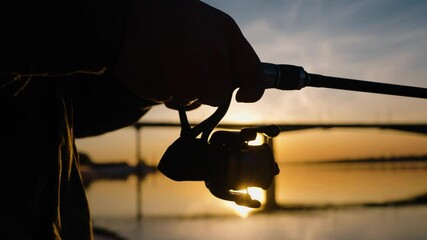 Fishing rod silhouette reeling line during sunset, capturing golden hour's serene moment with vibrant orange sky and water reflections, showcasing peaceful outdoor leisure activity