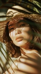 Young Asian woman in a light sun hat, sunlight streaming through palm leaves illuminating her serene expression.