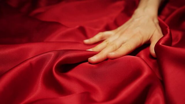 Woman's hand gently sliding and caressing luxurious red silk fabric, creating sensual, elegant movements with soft, flowing texture and delicate, intimate touch