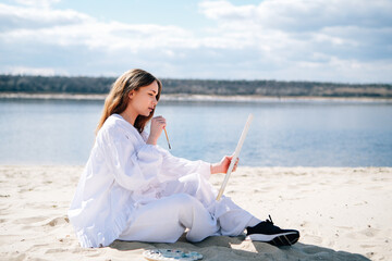 Cute teenage girl painting a picture on the river bank on a sunny day