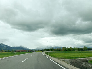 Winding road stretches through green fields toward snow-capped mountains under heavy overcast sky.