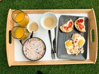Top view of breakfast tray on green grass with cereal bowl, coffee, orange juice, fried eggs, and bread with cheese and tomato spread.