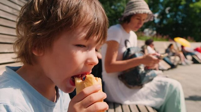 A little boy with blond hair sits on a bench outside in the park and eats a hot dog while his mom sits on the phone and watches social networks. Close-up view slow motion