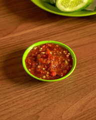 Spicy Red Sambal Made From Fresh Chilies And Shallots Served In A Small Green Bowl On A Wooden Table With Sliced Cucumber In The Background.