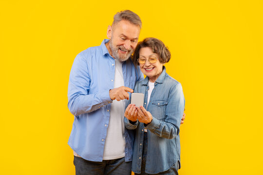 Happy senior couple looking at smartphone screen, using mobile app, shopping or banking online over yellow background