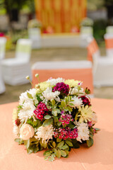 Floral centerpiece at an event, with white, yellow, and burgundy blooms atop a peach-colored surface. Chairs & stage visible in the background.