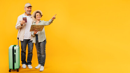 Excited retired couple tourists going vacation, posing with map, suitcase and flight tickets, pointing at copy space over yellow background