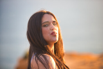 Fototapeta premium Young, Latin, brunette, beautiful woman smokes a cigarette and blows cigarette smoke in front of her. The woman stands on a cliff at sunset wearing blue underwear.