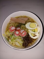 Bowl of Ramen with Assorted Toppings. A bowl of ramen with various toppings like boiled eggs, sausage, fish cakes, cabbage, and mushrooms, served on a textured surface