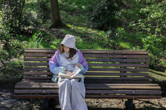 Young girl sits on a wooden park bench reading a book. She wears a casual outfit with a cap, enjoying a quiet outdoor moment. - Powered by Adobe