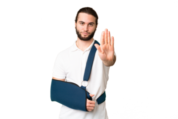Young handsome man with broken arm and wearing a sling over isolated chroma key background making stop gesture