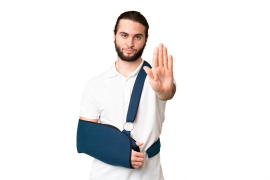 Young handsome man with broken arm and wearing a sling over isolated chroma key background making stop gesture
