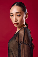 Fashion asian woman in sheer black top poses against red background with hair in ponytail and eyes...