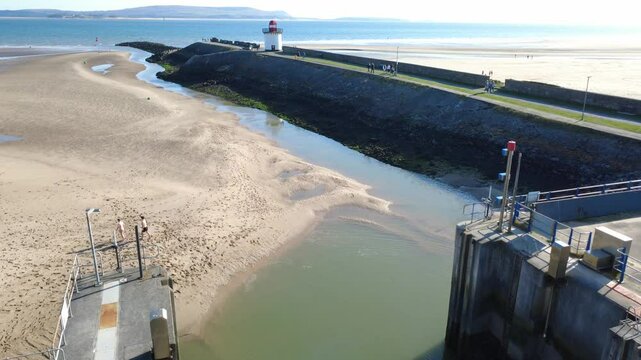 Drone shot travelling forwards from the entrance to Bury Port Harbour, along the breakwater, over the beach and past the lighthouse. Burry Port is in Caramarthenshire in South Wales, UK. A sunny sprin