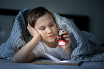 Cute kid boy teenager reading a book under the covers with flashlight