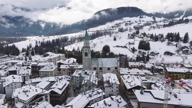 Aerial view of Cortina d'Ampezzo with its iconic bell tower, snow covered rooftops, alpine buildings, and misty Dolomite mountain slopes in winter.
