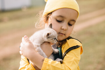 Adorable little girl in a yellow cap and raincoat holding kitty cat outdoor in nature. Pet concept.