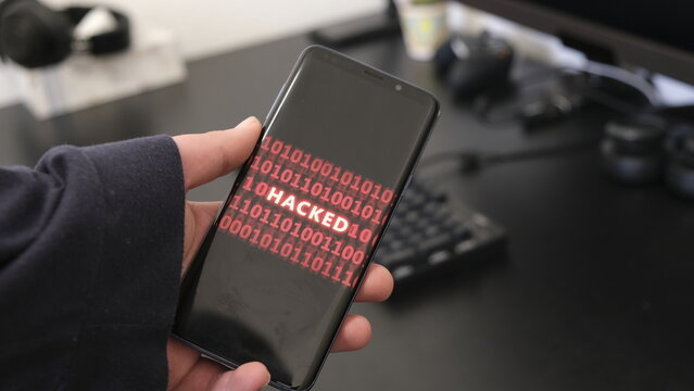 Smartphone screen with red binary code and "HACKED" in the center, held over a desk.