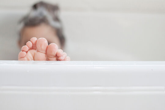 Girl sitting in a bath with shampoo in her hair and the focus on her bare feet sticking over the edge of the bathtub