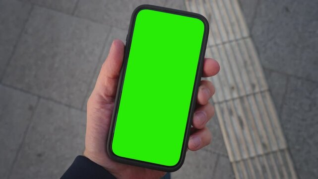 POV shot caucasian man looking at chroma key phone in his hand walking city street. Smart phone green screen in hand. Walk with cellphone, application mock up, camera locked, paving slabs background