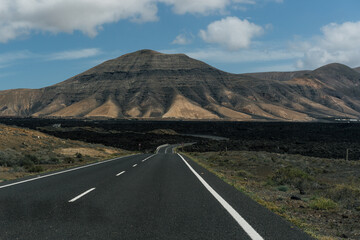 A road leading to volcano on a volcanic island, under a blue sky dotted with clouds. Lanzarote