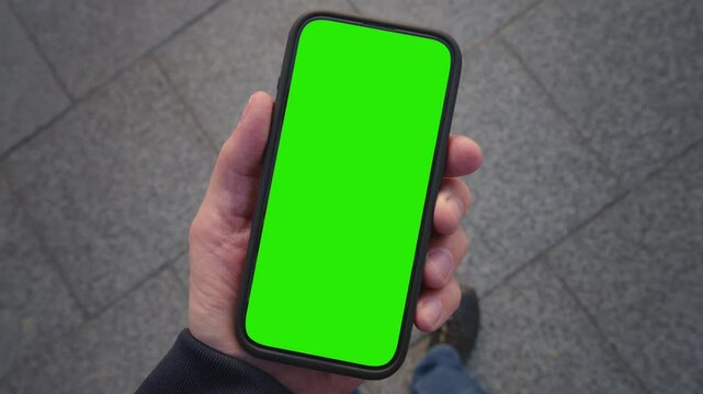 POV shot caucasian man looking at chroma key phone in his hand walking city street. Smart phone green screen in hand. Walk with cellphone, application mock up, camera locked, paving slabs background