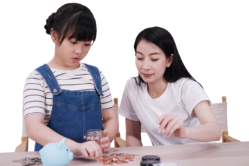 Smiling Asian daughter and mother putting coins into piggy bank for saving money for the future on wooden table. Child educational for homeschooling concept