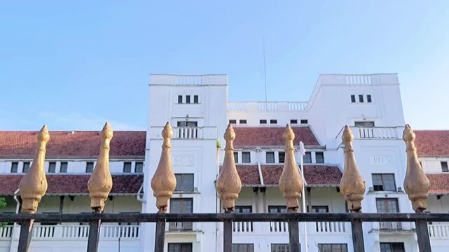Cinematic shot. A decorative iron fence with golden coated spears with the background of an old Dutch colonial building.