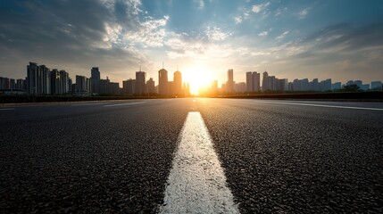 Obraz premium Wide asphalt road with double white lines leading to a city skyline of high-rise buildings under a sunset, warm golden-hour light filtering through clouds.