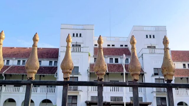 Cinematic shot. A decorative iron fence with golden coated spears with the background of an old Dutch colonial building.
