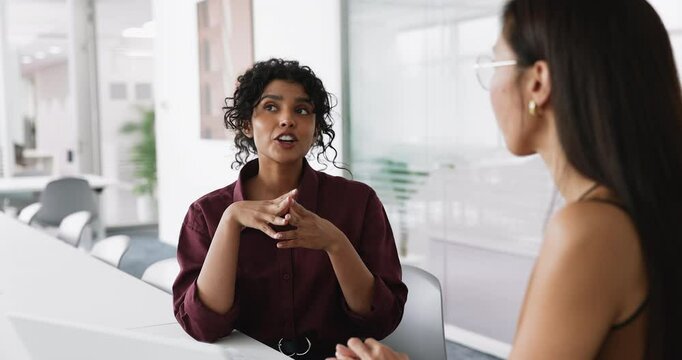 Young Indian woman speaking with female colleague during meeting seated at desk in modern office, applicant passing job interview, answers questions about education, professional skills and experience