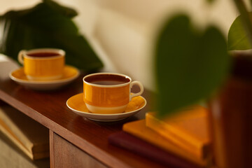 A pair of yellow vintage tea cups standing on a wooden table in a retro looking room framed by plants and books.